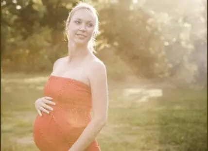 Pregnant woman in orange dress holding belly outdoors in natural sunlight
