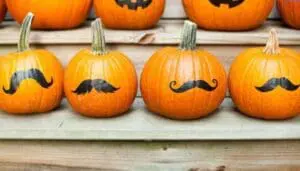 Four orange pumpkins decorated with black mustaches on wooden shelf