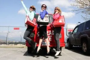 Three women holding large red reusable shopping bags while posing on a red car.
