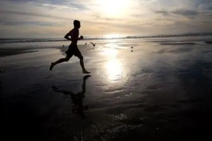 Silhouetted runner jogging along a wet beach at sunset with birds flying overhead