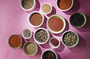 Assorted dried herbs and spices in white bowls on a pink background