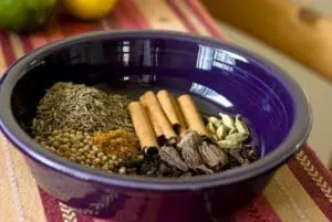 Indian spices in a purple bowl including cinnamon sticks, cardamom, and coriander seeds