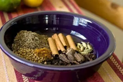 Indian spices in a purple bowl including cinnamon sticks, cardamom, and coriander seeds
