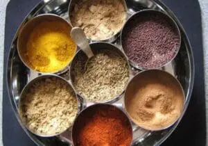 Seven spices and seasonings in metal bowls arranged on a tray, including turmeric, cumin seeds, and