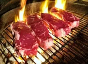 Red meat steaks grilling over open flames on a barbecue grate