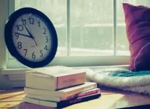 A cozy windowsill with a clock, stacked books, and a purple pillow in natural sunlight.