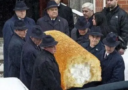 Pallbearers in dark coats carrying a large golden Twinkie cake during a funeral ceremony