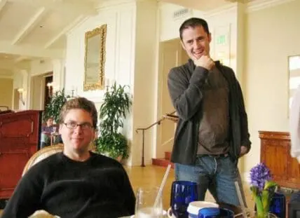 Two men in a casual indoor setting, one seated and one standing.