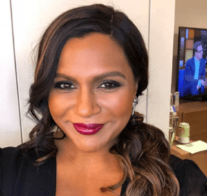 Mindy Kaling smiling at the camera wearing bold pink lipstick and a black blazer
