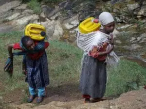 Two women carrying yellow water containers on their heads in a rocky terrain