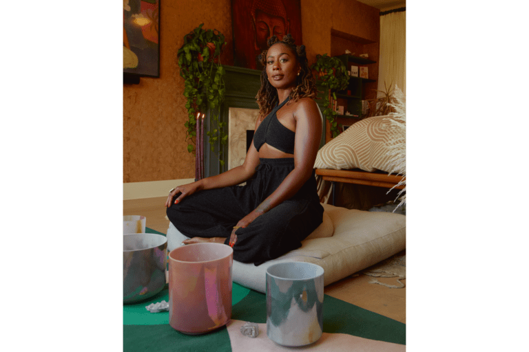 Image of actress Zuri Adele sitting in a relaxed, cross-legged meditation position on a floor pillow with candles and crystals in the foreground and a fireplace, plants, and artwork in the background showing elements of her personal wellness curriculum.