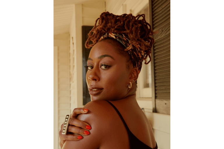 Image of actress Zuri Adele looking over her shoulder and holding onto her left shoulder with her right hand, brightly manicured and a unique bar ring on a porch with white siding and black shutters, invoking personal wellness.