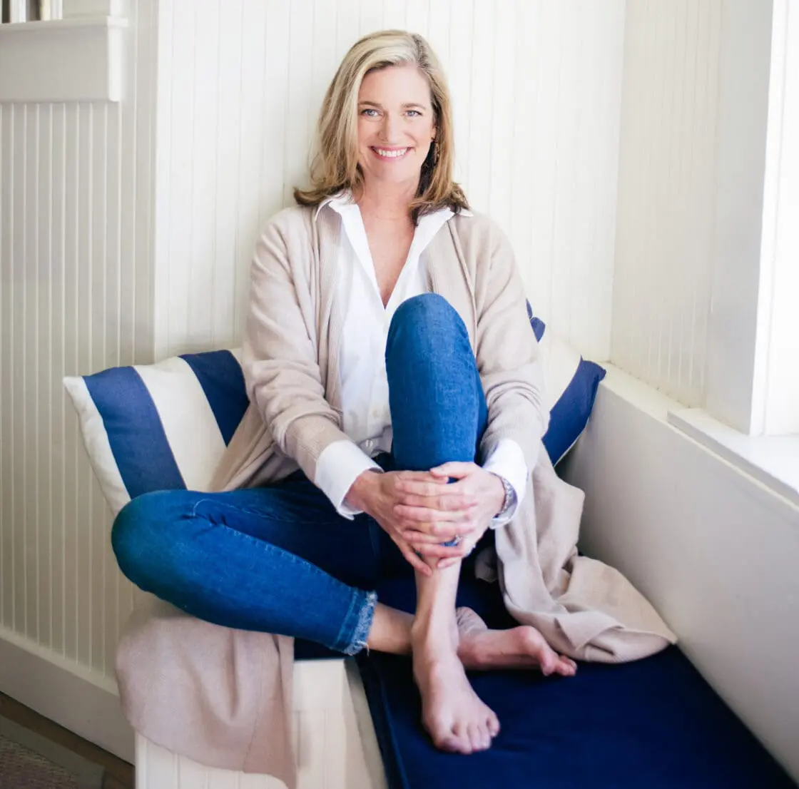 Smiling blonde woman in a cardigan and jeans sitting cross-legged on a window seat