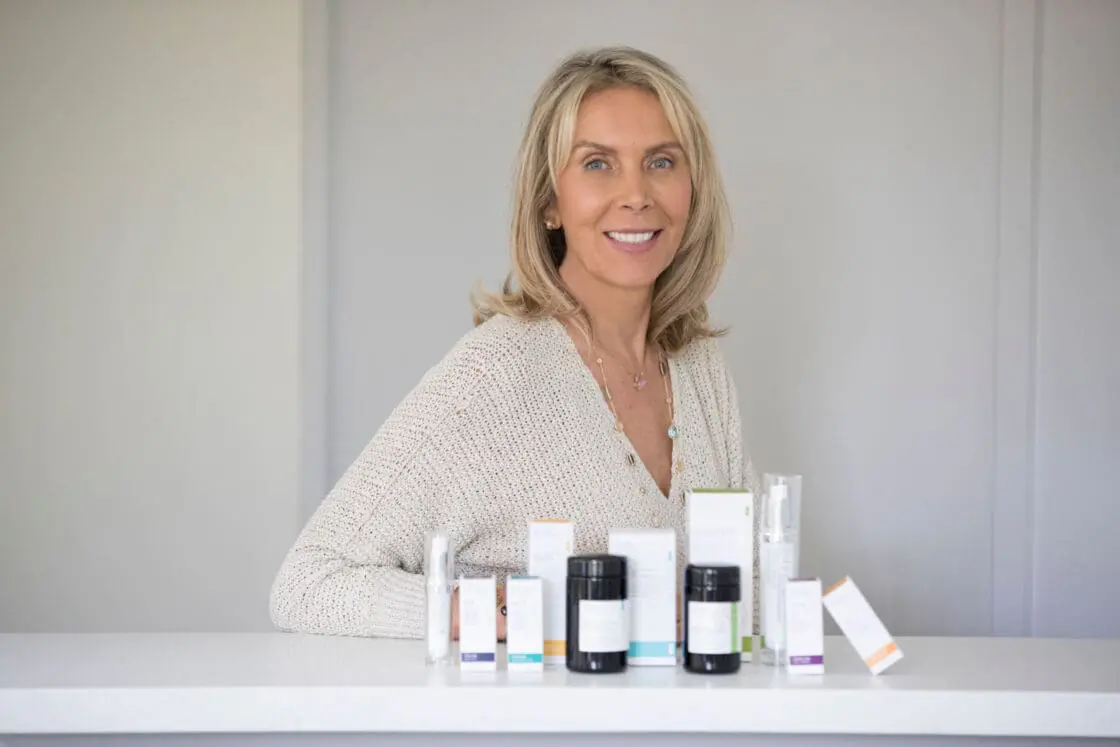 Smiling blonde woman in a beige sweater behind a counter displaying skincare product bottles