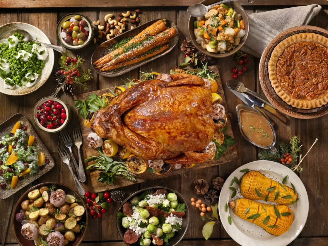 Image of an overhead view of thanksgiving dinner spread on a wood table with turkey, pecan pie, squash, roasted potatoes, salad, and cranberries.