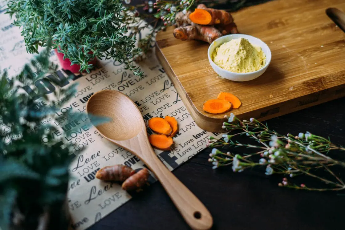 image of a wooden cutting board with a small white bowl containing ground ginger, with whole turmeric root sliced. Next to the board, which sits on top of a paper reading "Love, love, love" is a wooden spoon and more turmeric root. there are fresh herbs for Ayurvedic cleanse.