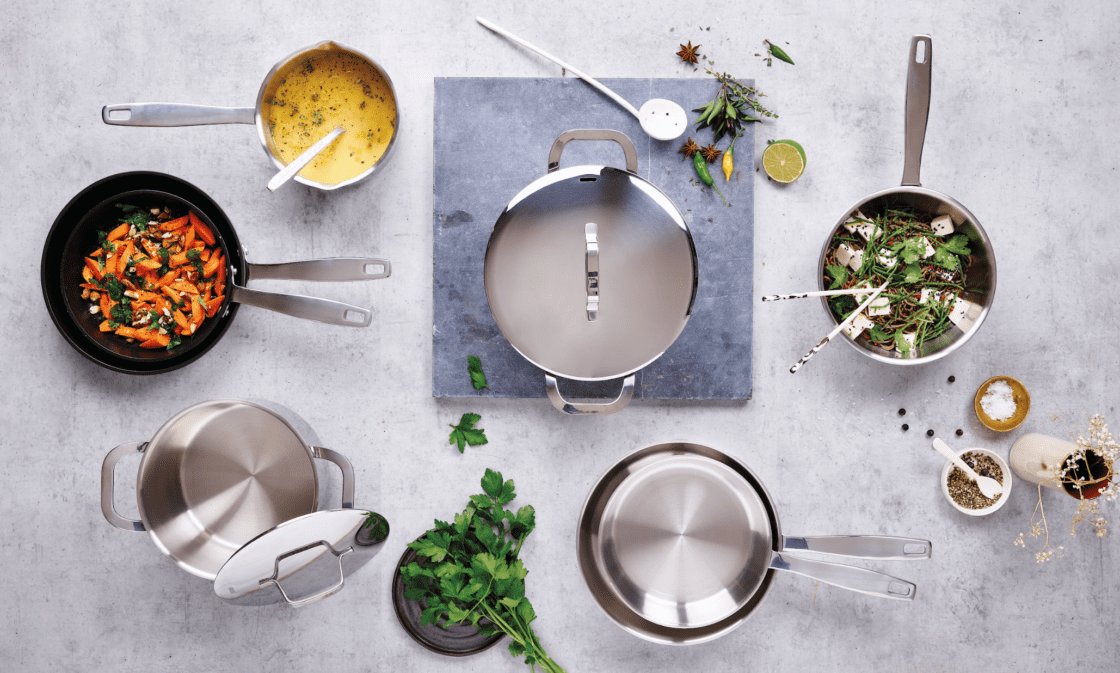 Image of Alva non-toxic cookware in many options from overhead view, stainless steel and ceramic coated nonstick with sauteed carrots, another pan with a tofu dish, and one with a buttery sauce with flecks of green and a spoon. Alva makes cookware from many different materials, they even have a cast iron skillet. Our chef tester Laura particularly loves the fry pan.
