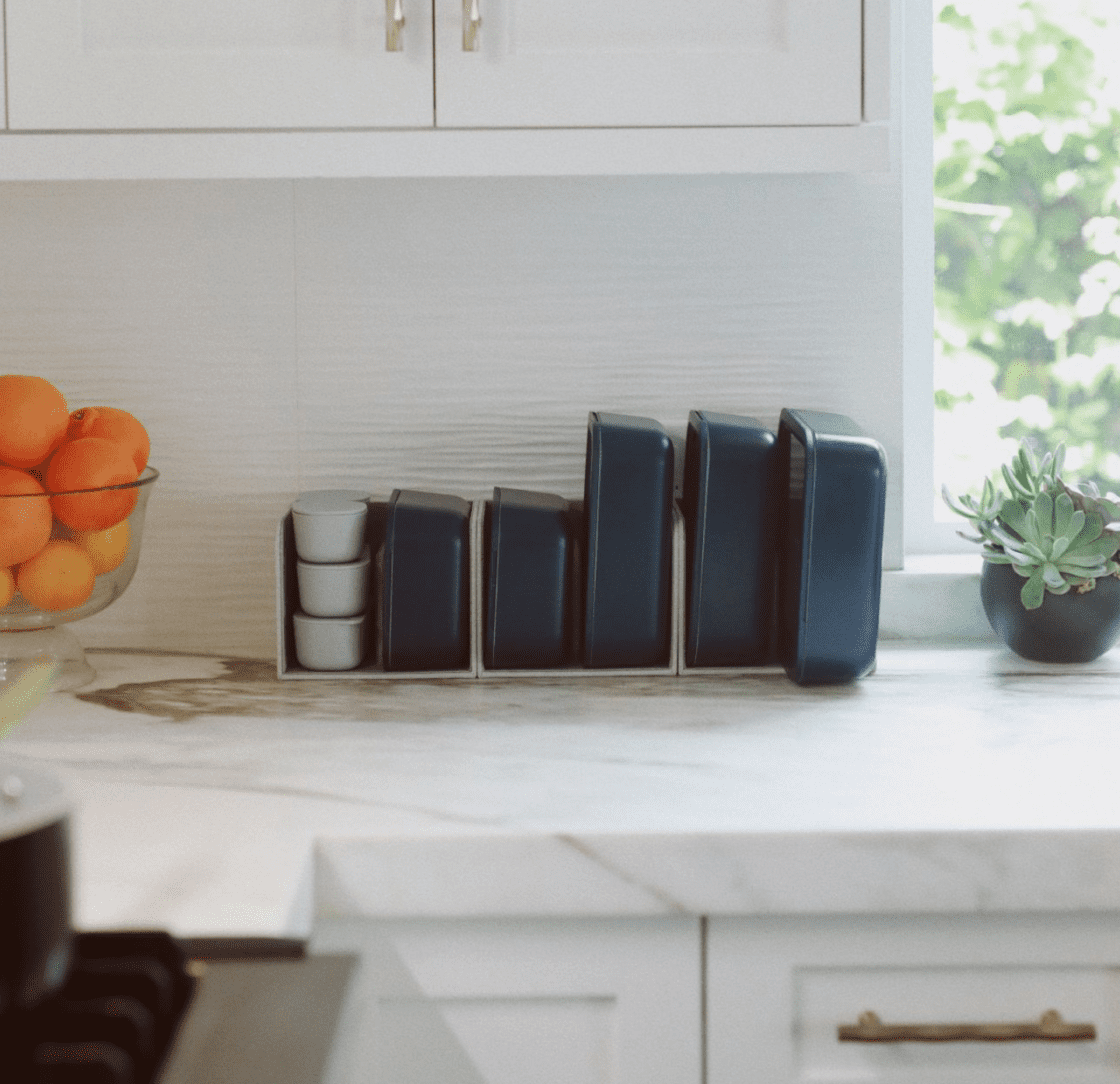 Image of Caraway Food Storage Containers in Navy, a full set in their storage organizer on a white marble counter with white cabinetry above next to a bright window and small succulent.