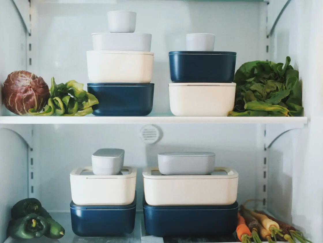 Image of Caraway Food Storage Containers in navy and cream in Laura Klein's refrigerator, two shelves shown with fresh greens, carrots, peppers, and purple cabbage.