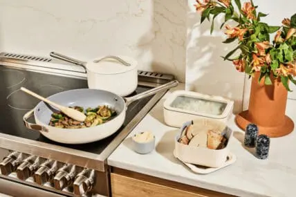 Image of Caraway Food Storage in cream, on a white marble countertop, prepped alongside a flat stovetop with amCaraway Home cookware Fry Pan and a covered saucepan to the rear. There's also a terracotta vase with fresh lilies, black marble salt and pepper shakers, and a veined marble backsplash.