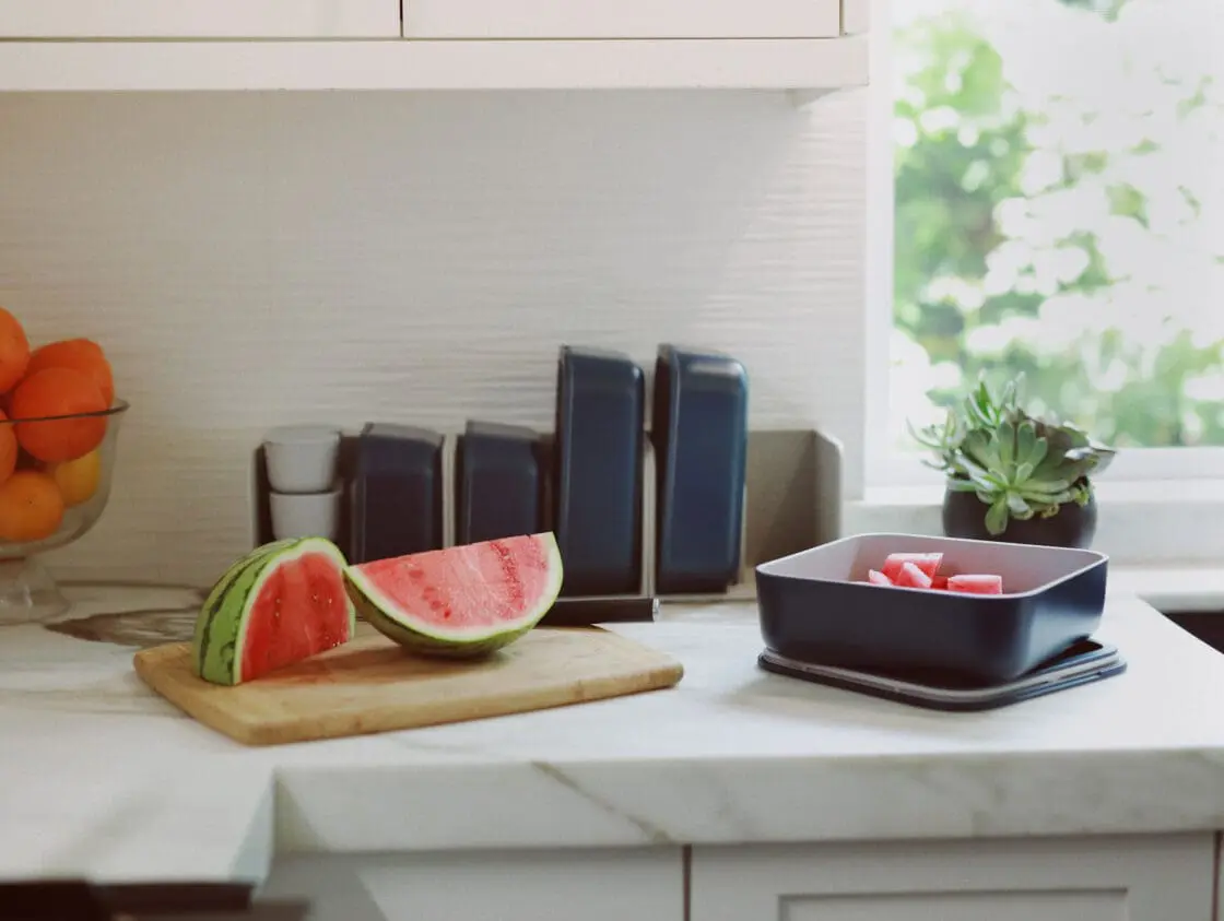 Image of Caraway Food Storage set stored in organizer on a counter in navy, apart from one container which sits out filled with cut watermelon on its lid. Next to it on the white marble counter is a small wood cutting board with watermelon and a glass bowl of clementines to the back right.
