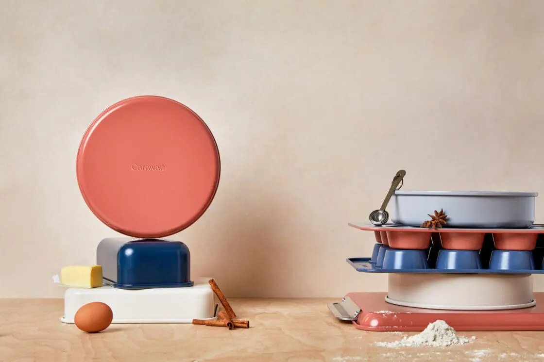 Image of Caraway Bakeware Set in mixed colors (perracotta, navy, and white) interestingly stacked in two stacks with a single brown egg, half stick of butter, and sprinkle of flour on the wooden work surface.