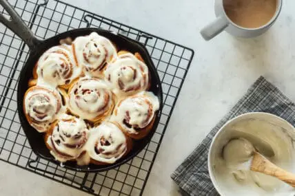 image of a Homemade vegan cinnamon roll recipe or buns baked in a cast iron skillet and covered with cream cheese icing. The skillet is cooling atop a black wire cooling rack on a white and gray marble countertop. A mug of coffee or tea is on the side next to a bowl full of icing.