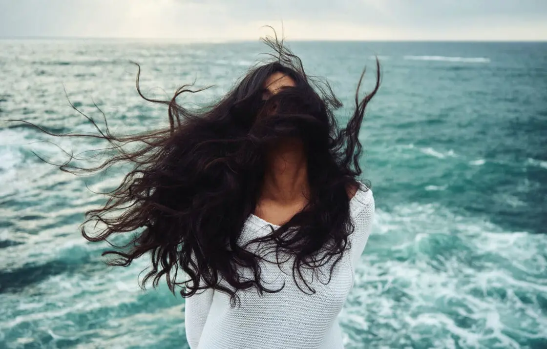Woman in the wind in front of the ocean with long hair blowing that is healthy because she supplements with marine collagen powder.