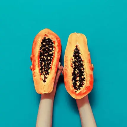 Image of cut open papaya held up on turquoise wall by two hands. Papaya is an excellent for supporting digestive enzymes