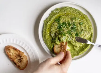 hand holding toast dipping into Fava Bean Pesto