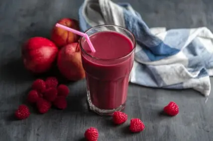 image of a clear glass of deep red liquid with a pink straw, surrounded by spilled raspberries and apples with a white and blue towel in the background for choosing a cleanse.