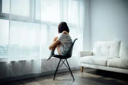 image of pensive woman sitting in chair looking out window through sheer, white curtains, next to white sofa wondering if she needs a full body cleanse.