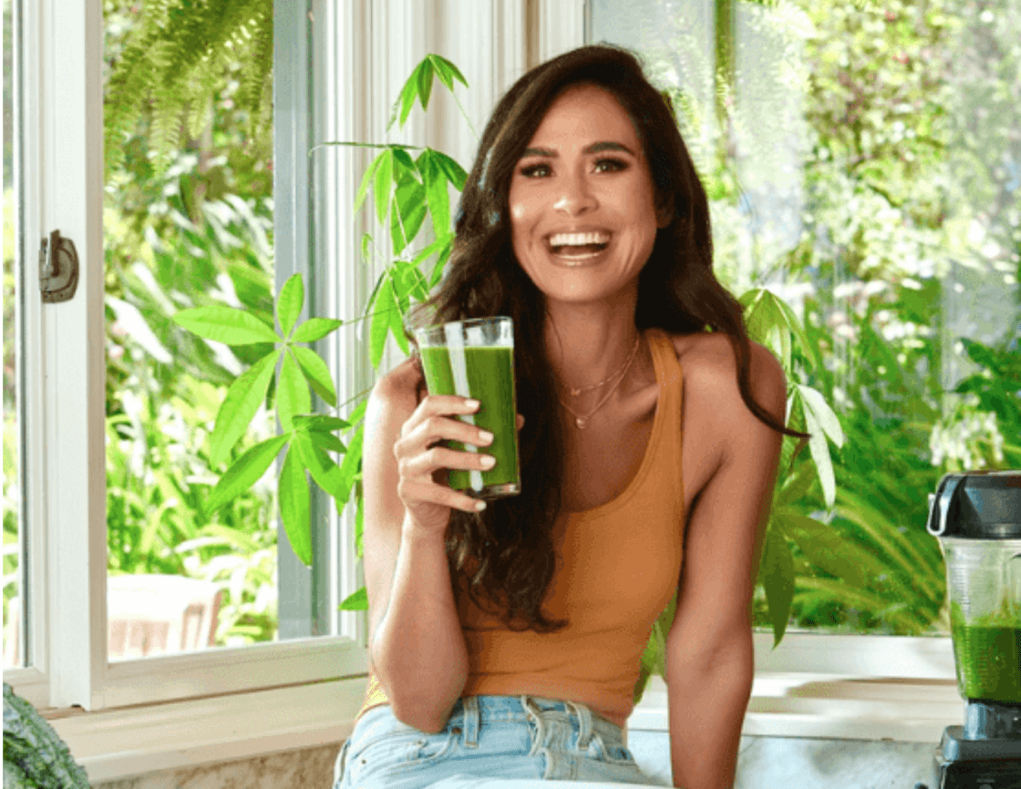 image of Kimberly Snyder sitting on kitchen counter next holding a large glass filled with a green smoothie. She wears an orange tank and light blue jeans, sitting next to a blender full of green smoothie and in front of a plant and window with green plants showing her whole body detox routine via Organic Authority.