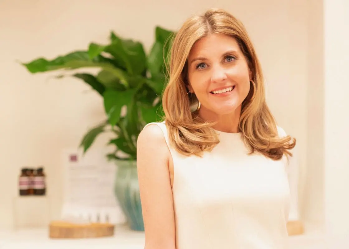 Smiling blonde woman in a white sleeveless top beside a leafy green plant