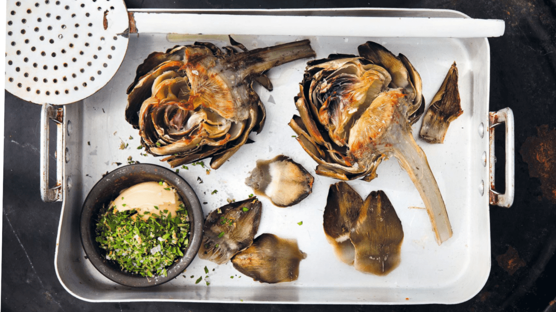 Image of grilled artichokes recipe in a white roasting pan with a black background and a small black ramekin of gremolata nestled in the corner, a white spider (large slotted spoon) rests on top of the roasting pan.