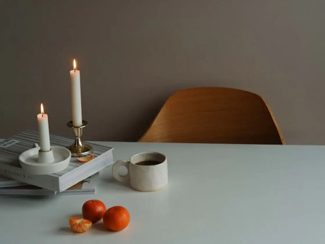 two tall natural candles burn above books on a table with a cup of coffee and three mandarin oranges.