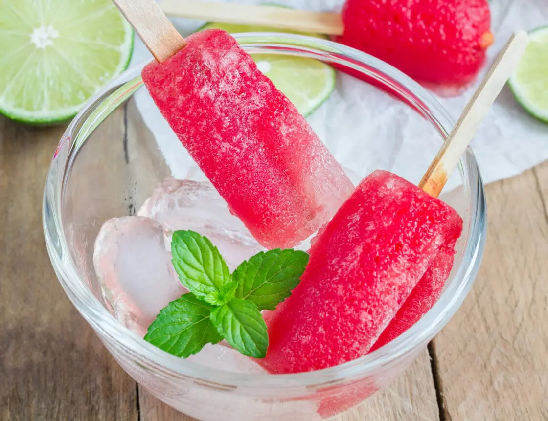 Image of two deep pink boozy popsicles made from vodka popsicle recipe over a clear glass bowl of ice served with a sprig of mint with sliced lime in the background via Organic Authority.