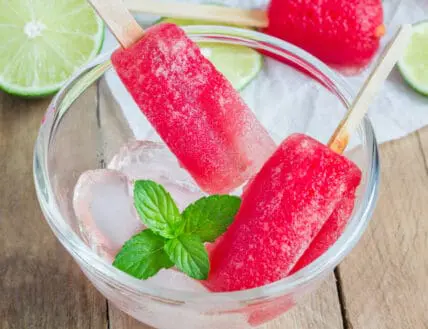 Image of two deep pink boozy popsicles made from vodka popsicle recipe over a clear glass bowl of ice served with a sprig of mint with sliced lime in the background via Organic Authority.