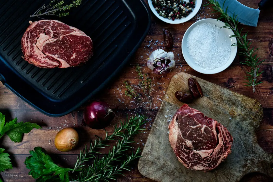 two cuts of steak, one on a wood cutting board and one on a grill with aromatic ingredients scattered around the counter, humanely raised meat via Organic Authority