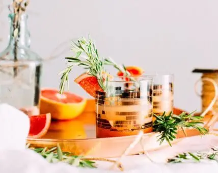 Image of a cocktail tumbler with gold accents and with squeeze of grapefruit on the rim with a rosemary sprig in the glass. sprigs of rosemary are scattered around, and there is a cut grapefruit in the background, along with a non-alcoholic spirits decanter.