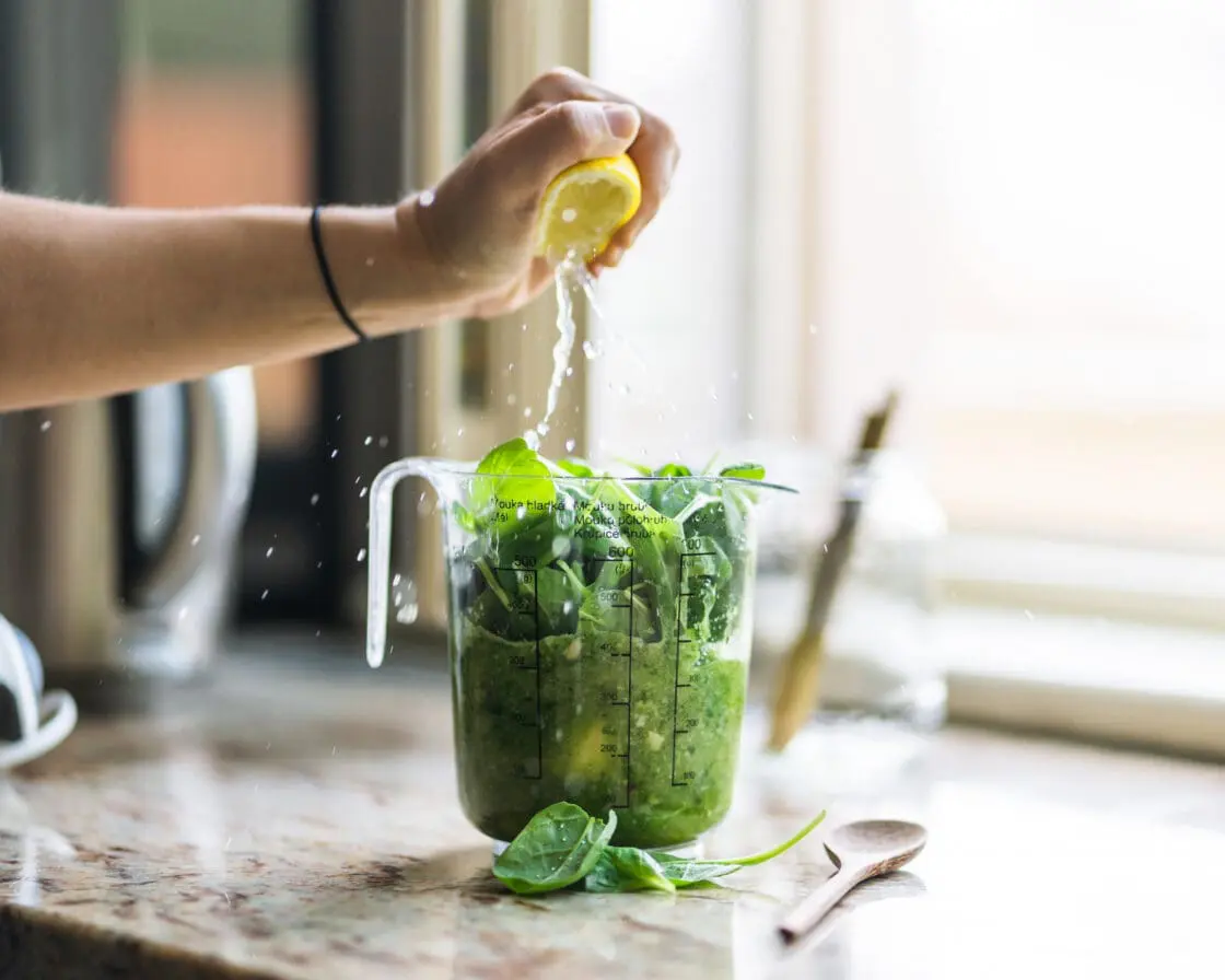 image of a blender with greens and lemon being squeezed into it on a counter with a wooden spoon and a few loose leaves in front a an out of focus window. Which cleanse is right for you?