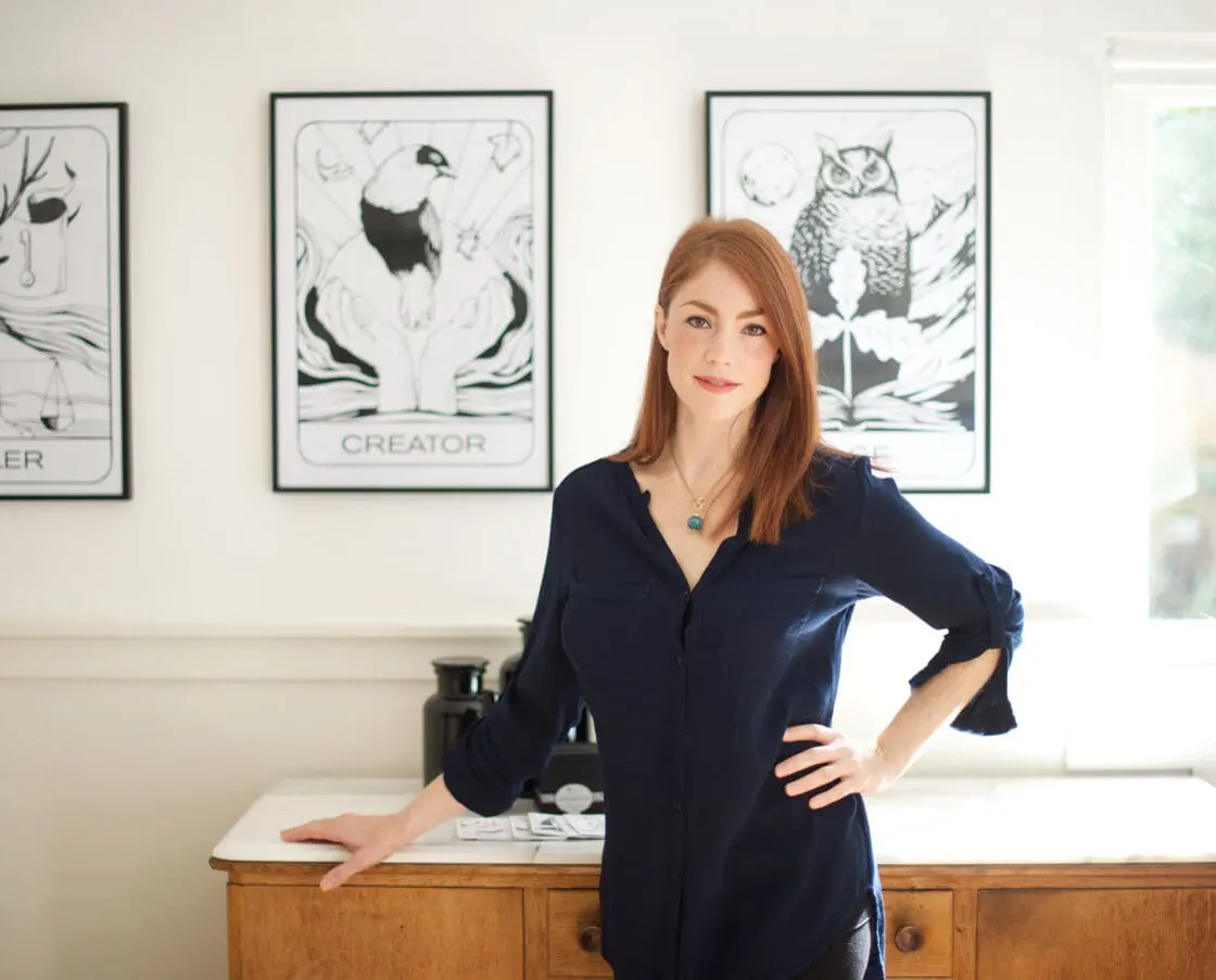 Red-haired woman in a navy blouse standing by a cabinet with framed line-art prints behind her