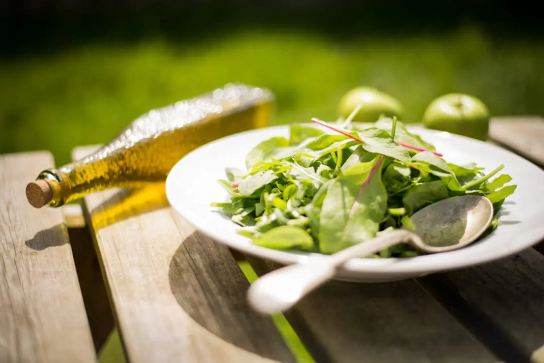 image of white bowl of leafy greens with worn spoon on a wooden table outside with a bottle of olive oil beyond the bowl showing healthful, ingredients for detoxifying the liver.