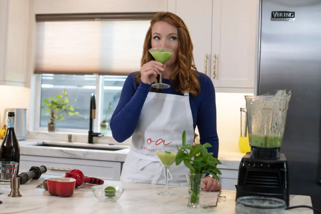 image of Laura Klein sipping cold melon soup from a martini glass in her kitchen with Vitamix blender on the marble island filled with the green melon soup.
