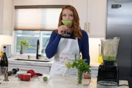 image of Laura Klein sipping cold melon soup from a martini glass in her kitchen with Vitamix blender on the marble island filled with the green melon soup.