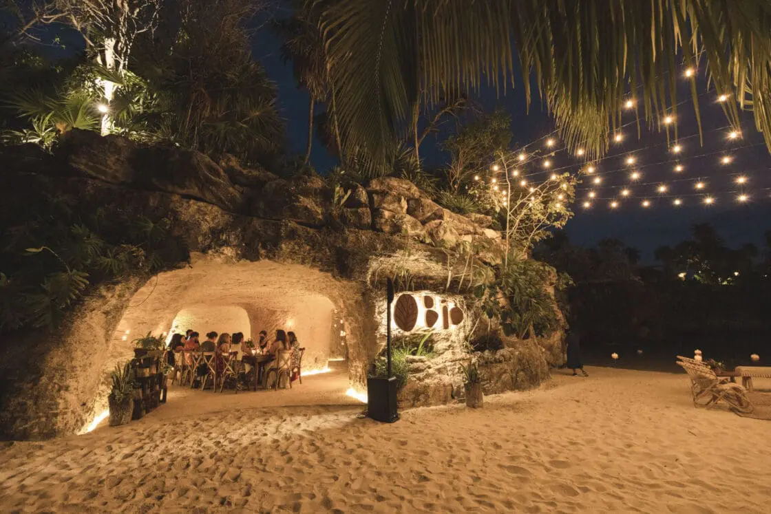 Image of a beach with a natural stone building beneath palms and open to the elements. Inside, you view a cozily lit dining situation and relaxed diners, as is to be expected of a wellness retreat on a beach in Mexico.