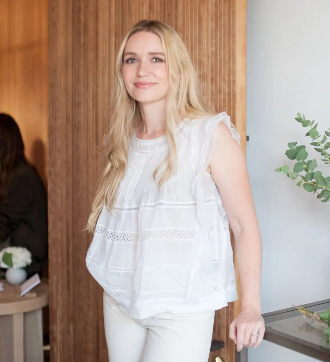Smiling blonde woman with long hair wearing a white sleeveless ruffled top in a wood-paneled room