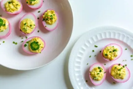 Image of pickled deviled eggs on two white plates. The eggs are pretty with a pink exterior, giving a lovely pink rim around the cut side, filled with yellow yolk filling and topped with chives and thinly sliced jalapenos.