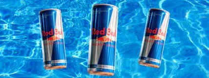 Image of 3 cans of Red Bull with a blue water background showcasing Red Bull ingredients but you'll want to know, are they safe to drink?