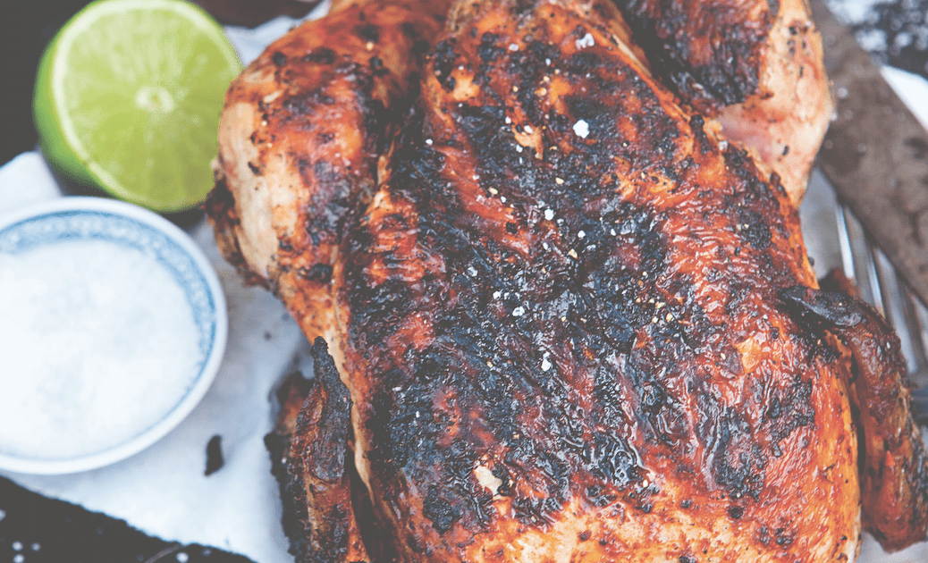 image: whole grilled chicken on a black enamel plate with a halved lime and sea salt in a small bowl and sprinkled around a bottle of Tabasco for whole grilled chicken recipe with chipotle rub via organic authority.